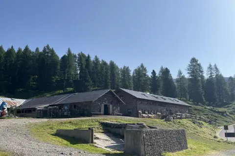 Malga Vallazza, un rifugio alpino in pietra con tetti scuri, circondato da un bosco di abeti sotto un cielo azzurro.