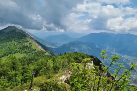 Panoramica mozzafiato dal Monte Stivo, con valli verdi e montagne azzurre sotto un cielo nuvoloso.