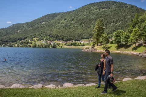 Coppia passeggia sul lungolago del lago delle piazze, con montagne verdi e un villaggio sullo sfondo.