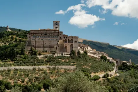La Basilica di San Francesco domina il colle di Assisi, circondata da uliveti e montagne verdi sotto un cielo azzurro con nuvole bianche.