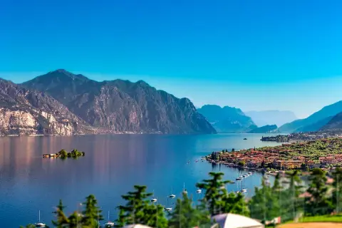 Un incantevole panorama per le tue vacanze sul Lago di Garda: montagne maestose, un'isola verde e un borgo pittoresco con barche a vela.