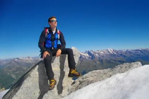 Giovane alpinista seduto sulla cima Presanella, ammira il panorama mozzafiato delle Alpi sotto un cielo azzurro.