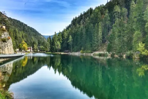 Un **lago smeraldo** incastonato tra montagne boscose, con un ponte pedonale che riflette le sue acque cristalline.