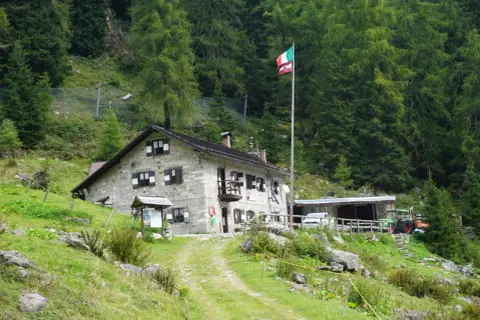 Il rifugio Trivena, con bandiera italiana, è una casa in pietra immersa nel verde, con un trattore parcheggiato accanto.
