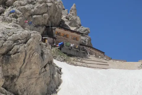 Rifugio Passo Principe, un rifugio alpino in legno incastonato tra rocce imponenti e neve, con un cielo azzurro limpido.