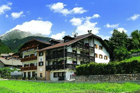 Albergo La Montanara a Predazzo, con le sue balconate in legno e le montagne sullo sfondo, incorniciato da un cielo azzurro e nuvole bianche.