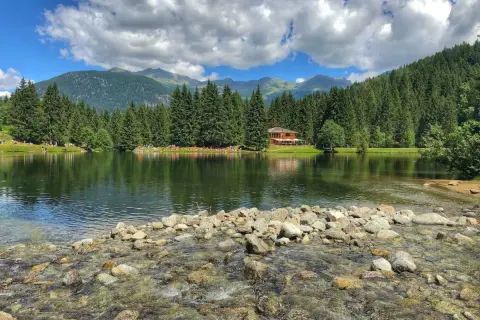 Il lago dei caprioli, circondato da fitti boschi e montagne, riflette un cielo azzurro con nuvole bianche. Una casa in legno si affaccia sull'acqua.