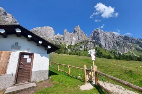 Malga Cavallera: sentiero con cartelli direzionali e rifugio bianco ai piedi delle maestose montagne.