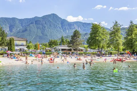 Spiaggia affollata al camping Riviera Caldonazzo, con gente che prende il sole e nuota nel lago, montagne sullo sfondo.