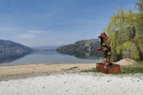 Scultura metallica di un cane sulla riva del lago di Caldonazzo, con montagne riflesse sull'acqua calma.