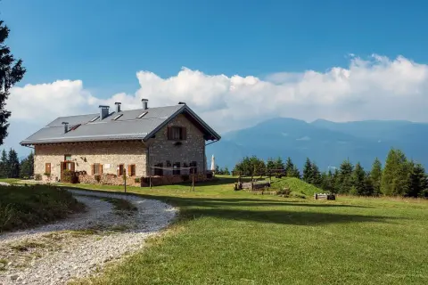 Malga Campo, un rifugio in pietra con tetto metallico, immerso nel verde di un prato alpino, con montagne sullo sfondo.