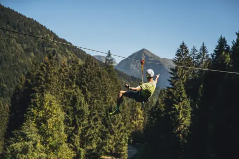 Uomo in volo su una zipline in un parco avventura Trentino, circondato da fitti boschi e montagne.