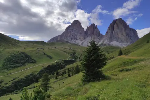 Verdi pendii alpini verso le imponenti cime del Sassolungo, con sentieri e impianti di risalita. Immagina le cascate basaltiche di Canazei in lontananza.