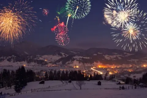 Fuochi d'artificio colorati illuminano il cielo notturno sopra un villaggio innevato, celebrando il capodanno in montagna.