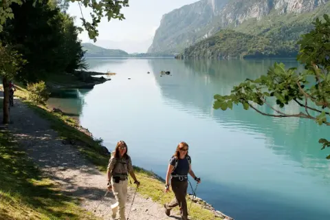 Due escursioniste camminano lungo il sentiero del lago di Molveno in estate, con montagne verdi sullo sfondo.