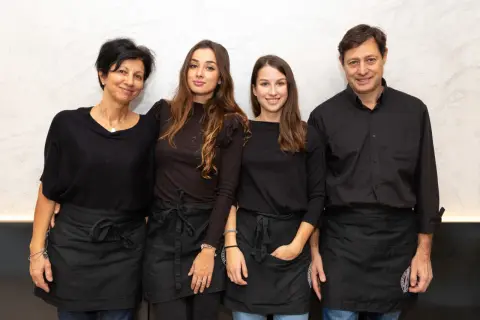 Four people, two women and two men, wearing black shirts and aprons, stand together smiling.