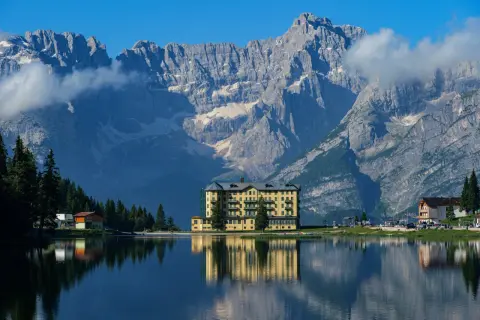 Un hotel giallo si specchia in un lago alpino, con imponenti montagne sullo sfondo: ecco dove si trovano le Dolomiti.