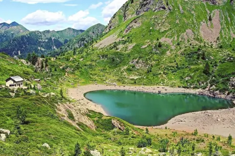 Un rifugio solitario vicino al **lago di Erdemolo**, circondato da verdi pendii montuosi e un cielo azzurro con nuvole bianche.