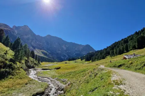 Sentiero sterrato che sale verso un rifugio alpino, con un ruscello che scorre in primo piano e montagne imponenti sullo sfondo, tipico del passo Duron.