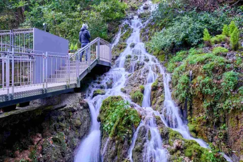 Una persona osserva le **cascate del Varone** da una passerella metallica. L'acqua scende a cascata su rocce ricoperte di muschio, immersa nel verde.