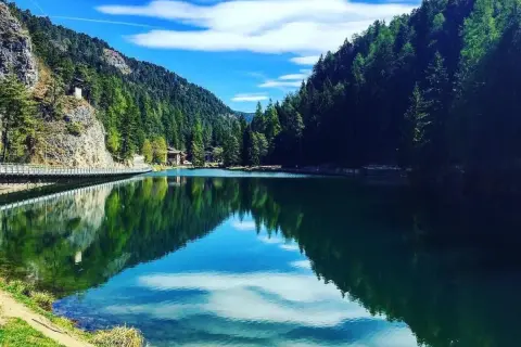 Un lago smeraldo incastonato tra montagne boscose. Una spiaggia erbosa invita al relax, mentre un ponte attraversa le acque calme.