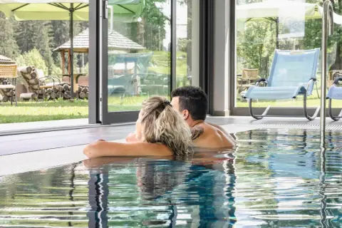 Coppia rilassata nella piscina interna di un wellness hotel Dolomia, con vista sul giardino e sulle montagne.