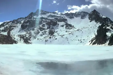 Il lago delle stellune, ghiacciato e circondato da montagne innevate, riflette il cielo azzurro e le nuvole.