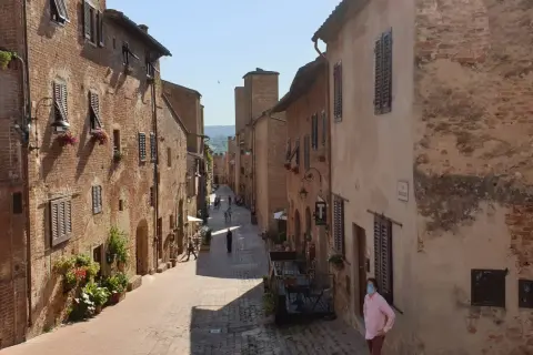 Una persona con mascherina passeggia per una stretta via acciottolata di **Certaldo Alto**, circondata da antichi edifici in mattoni con persiane in legno.