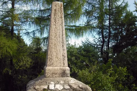 Obelisco in pietra sul **monte Zugna**, circondato da alberi di larice e vegetazione rigogliosa.