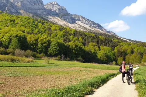 Ciclisti su un sentiero sterrato, con campi coltivati e un bosco rigoglioso ai piedi di una montagna imponente. Un passo bordala da godersi in bicicletta.