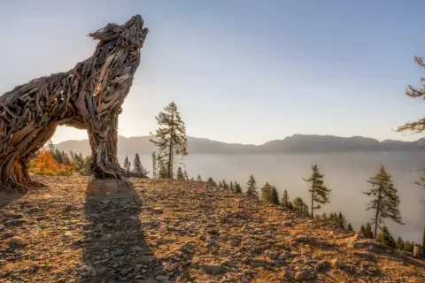 Scultura di una lupa del Lagorai in legno, che ulula al tramonto su un crinale roccioso, con montagne nebbiose all'orizzonte.