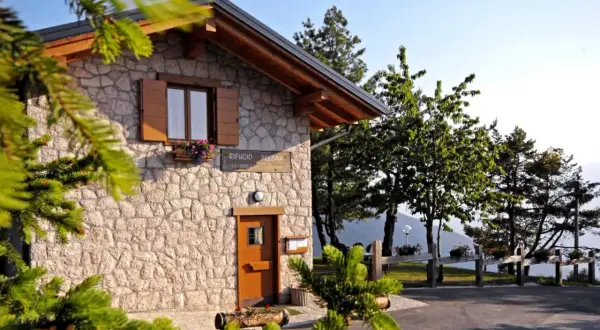Rifugio Predaia, un'accogliente costruzione in pietra con tetto spiovente, circondato da alberi verdi e un cielo azzurro.