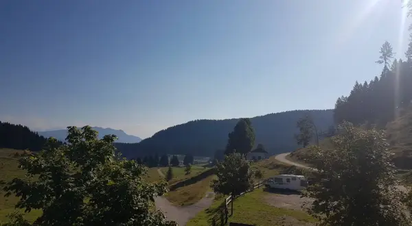 Un camper parcheggiato vicino a un **rifugio barricata** in montagna, con un sentiero che si snoda tra prati verdi e boschi.