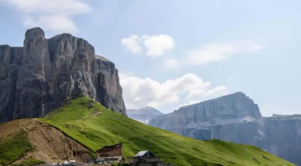 Edifici alpini e un parcheggio affollato ai piedi di imponenti pareti rocciose sul passo Sella, con verdi pendii erbosi.