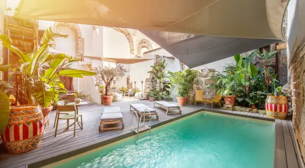 Courtyard pool with lounge chairs, lush plants, and a decorative barrel under a sunshade.