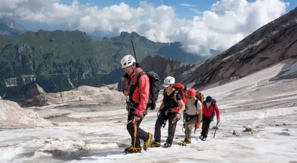 Alpinisti in cordata attraversano un ghiacciaio innevato, con montagne verdi sullo sfondo. Un'avventura al Passo Fedaia.