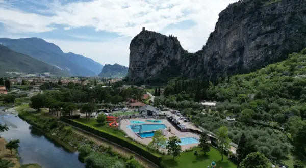 Agricamp Altogarda: un villaggio vacanze con piscine e scivoli ai piedi di imponenti montagne, vicino a un fiume e un borgo.