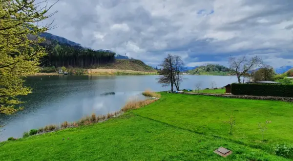 Un tranquillo lago di Pinè con verdi prati, alberi spogli e nuvole minacciose nel cielo.