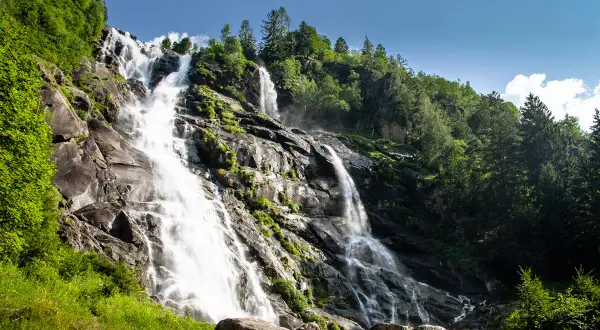 Cascate maestose nel Val di Genova, con acqua che scende impetuosa su rocce scure, circondate da fitta vegetazione verde e alberi imponenti sotto un cielo azzurro.
