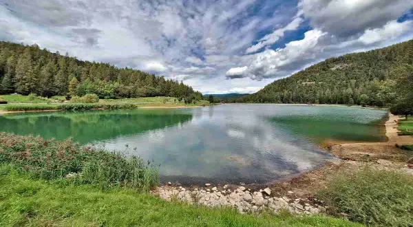 Il lago di Tavon, circondato da boschi verdi, riflette un cielo nuvoloso.