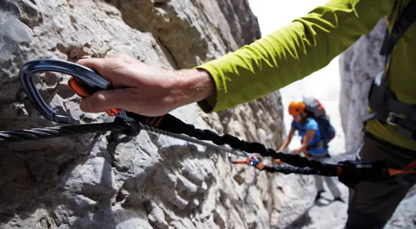 Mano agganciata a cavo d'acciaio su una parete rocciosa, un'avventura tra le ferrate Trentino con un'altra persona sullo sfondo.
