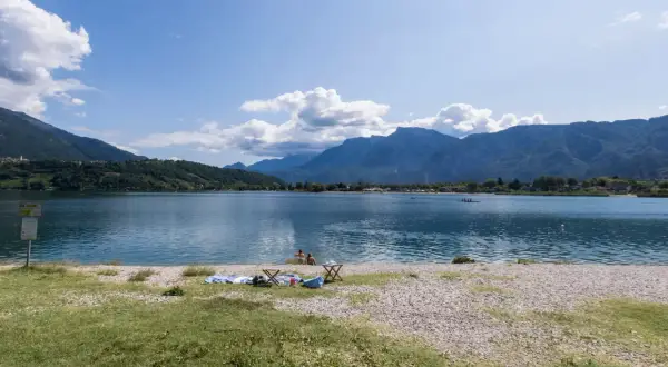 Relaxing day al lido di Caldonazzo, con montagne sullo sfondo e persone che si godono il sole e il lago.
