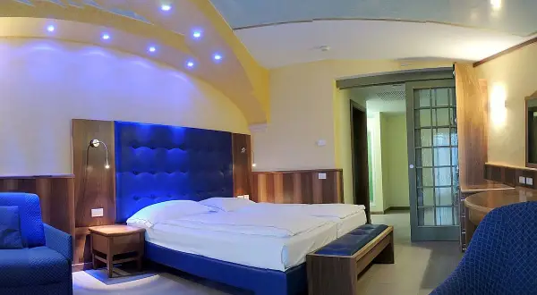Camera moderna con letto king size e testiera blu imbottita, illuminazione soffusa e soffitto decorato. Un assaggio del comfort del Grand Hotel Astoria Lavarone.