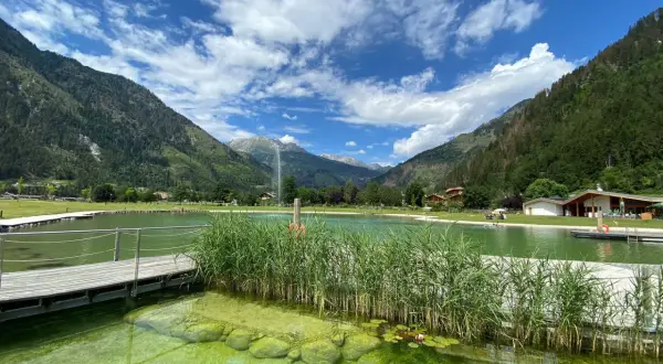 Un rinfrescante biolago a Predazzo, circondato da montagne verdi e un cielo azzurro con nuvole bianche. Pontili in legno e canneti creano un'atmosfera serena.
