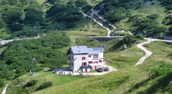 Il rifugio Scalorbi, un edificio in pietra con persiane rosse, si erge in una valle verdeggiante, circondato da sentieri e vegetazione alpina.