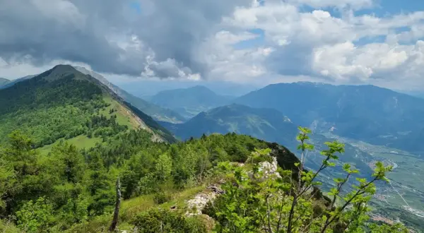 Panoramica mozzafiato dal Monte Stivo, con valli verdi e montagne azzurre sotto un cielo nuvoloso.
