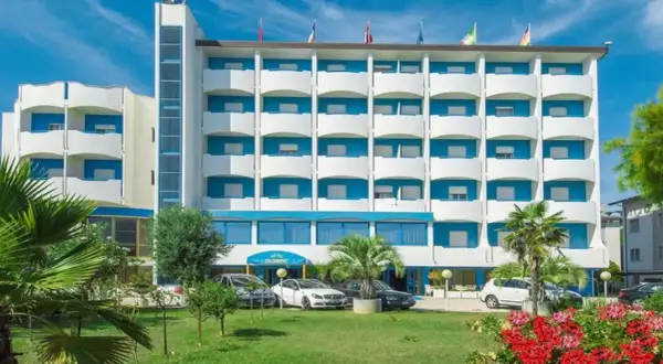 Hotel Olympic, un edificio bianco e blu con balconi, circondato da verde e fiori. Auto parcheggiate davanti.