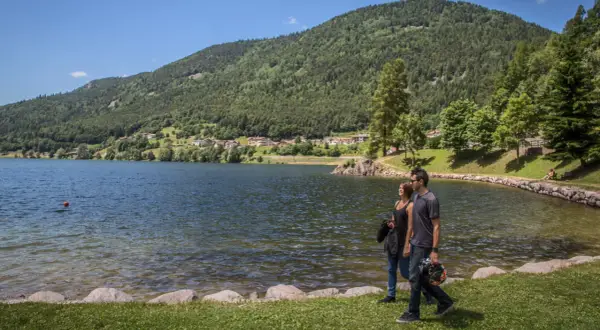 Coppia passeggia sul lungolago del lago delle piazze, con montagne verdi e un villaggio sullo sfondo.