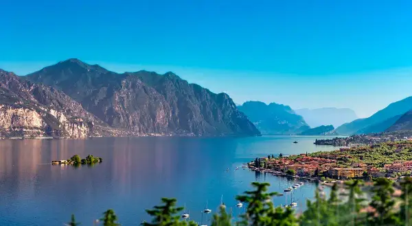 Un incantevole panorama per le tue vacanze sul Lago di Garda: montagne maestose, un'isola verde e un borgo pittoresco con barche a vela.