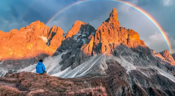 Persona seduta ammira le maestose Pale di San Martino illuminate dal sole al tramonto, con un arcobaleno che le sovrasta.